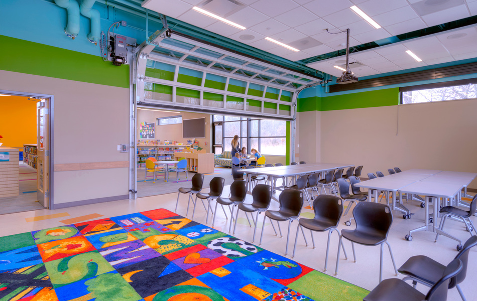 Interior of the Garage area of the Youth department with tables set up in rectangles and chairs around a colorful rug on the floor.