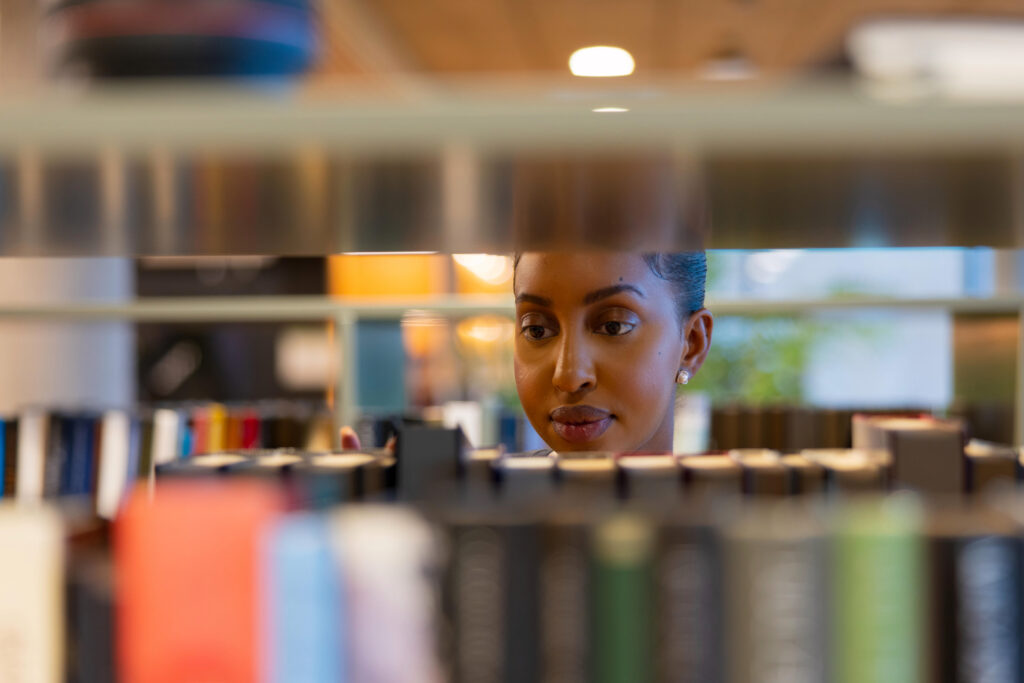 A focused individual is browsing through a library bookshelf, thoughtfully selecting books. The scene captures an atmosphere of learning and focus, evoking curiosity and ambition. The warm lighting and blurred background enhance the contemplative mood, symbolizing education and study in a peaceful environment.
