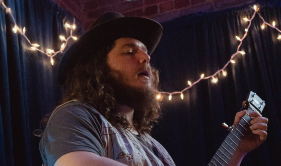Bearded man with a banjo singing in front of string lights