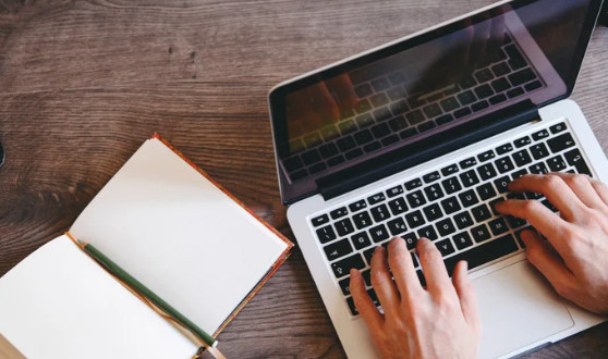 Person typing on a laptop with a notebook open beside them
